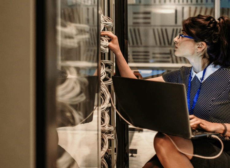 IT support technician troubleshooting network cables and server rack connections while working with a laptop.